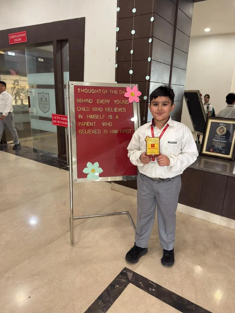 Young boy in school uniform holding an award certificate, standing beside an inspirational quote board about parental belief and confidence