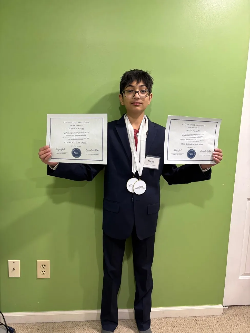Boy in a suit holding two certificates and wearing a medal, celebrating academic and leadership achievement at Oratrics